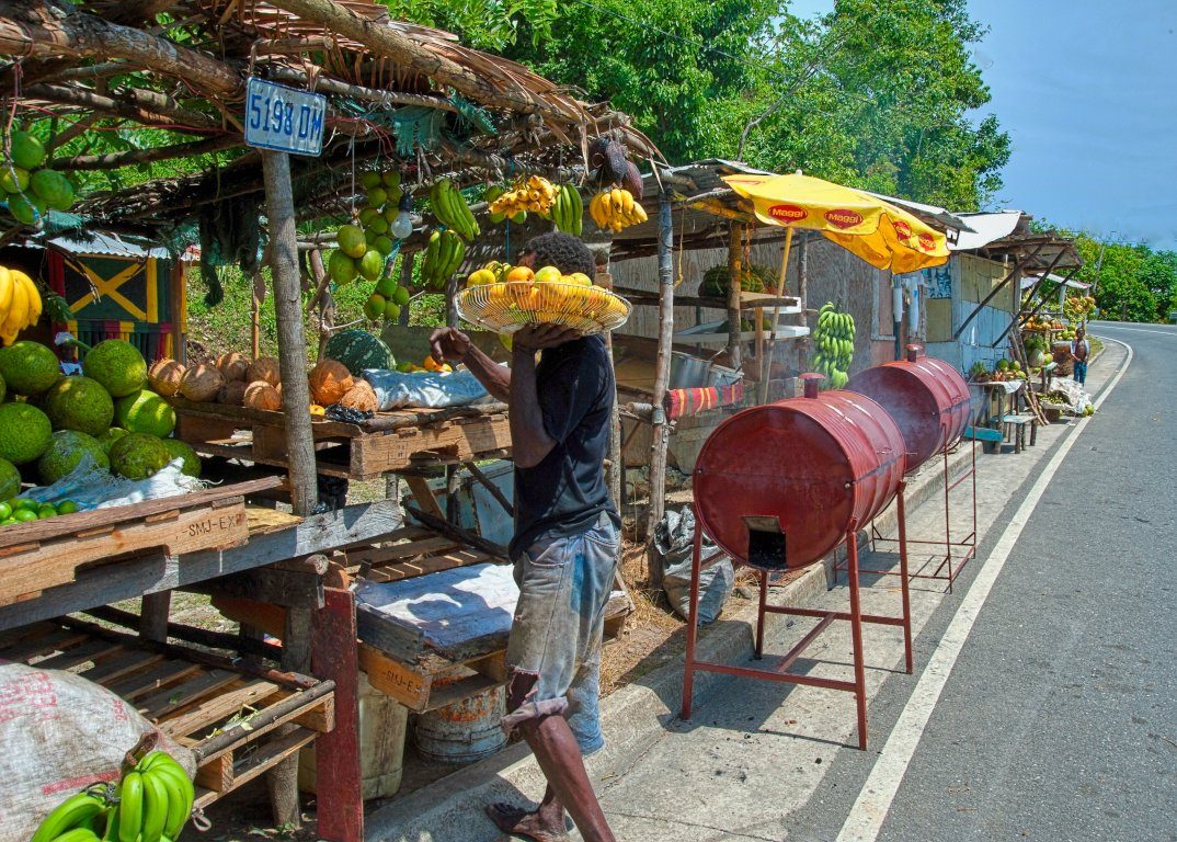 A man carries a basket of fruit past a roadside stall in Jamaica